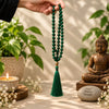 A hand holds a dark green beaded necklace with a tassel against a warm, sunlit background with a Buddha statue, potted plants, and a candle.