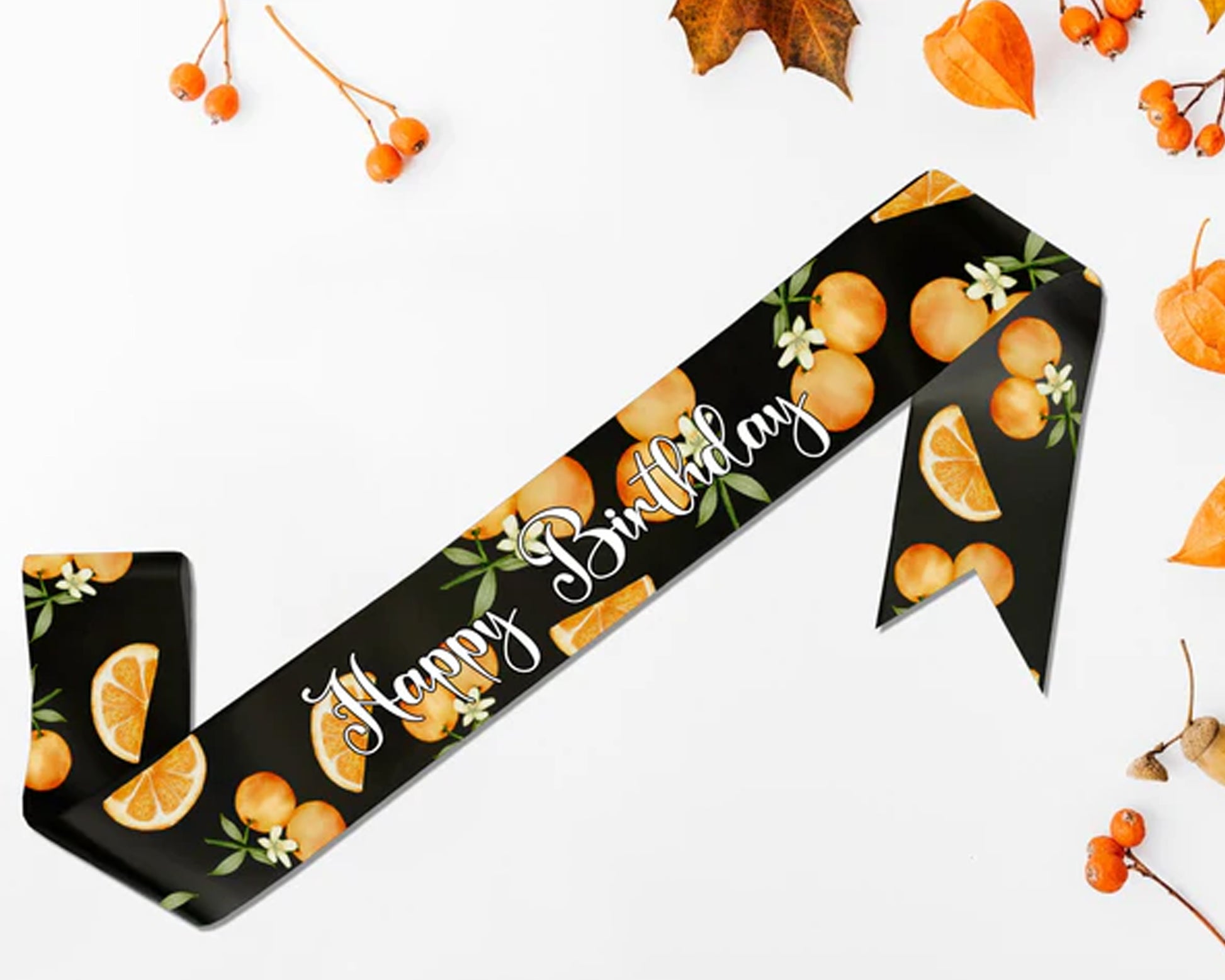 A "Happy Birthday" sash featuring a vibrant orange-themed design on a black background, surrounded by autumnal decorations.