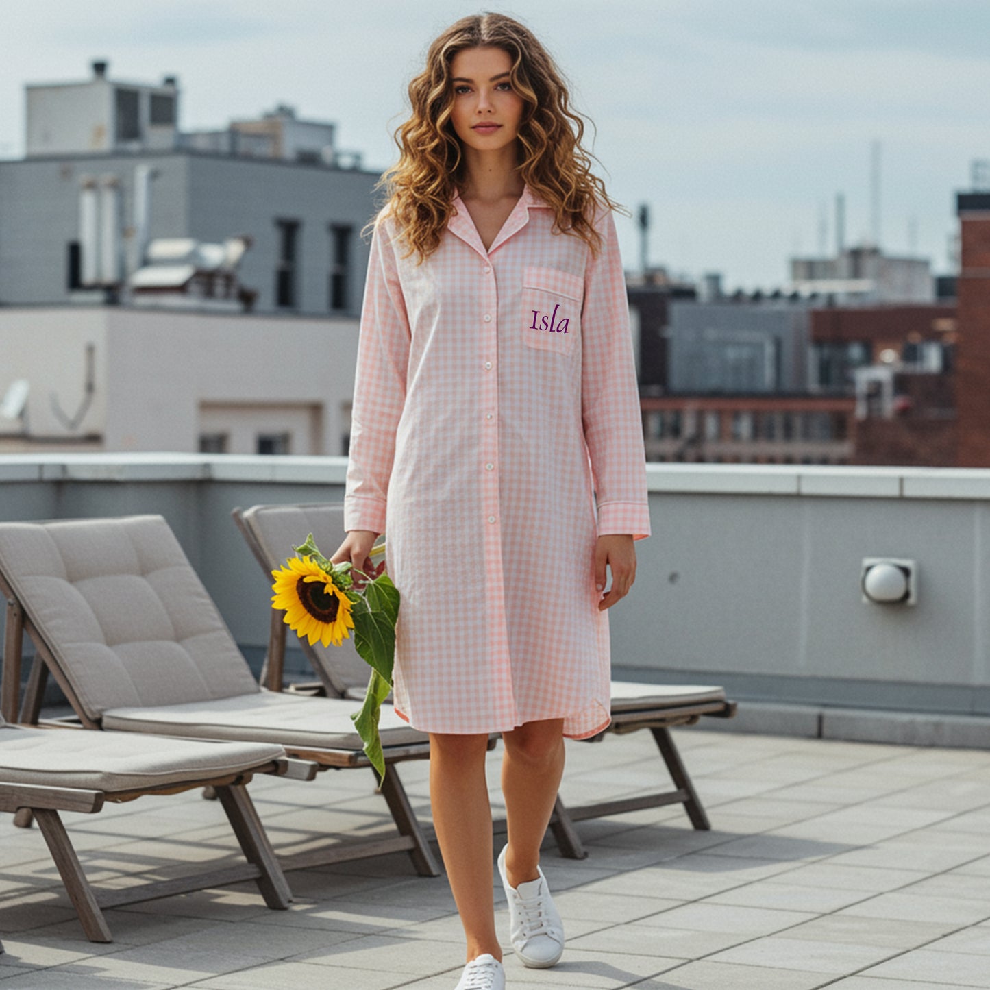 Woman walking on a terrace holding a sunflower wearing a pink gingham sleepshirt with Isla embroidered on the pocket