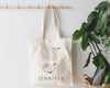 Cream-colored tote bag with the letter "J" decorated with delicate flowers and green leaves, with "JENNIFER" printed below, hanging on a wooden hook beside a lush green plant.