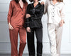 Three women wearing stylish satin pajama sets in black, bronze, and champagne, featuring button-up tops with contrast piping and matching pants.