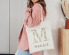 Stylish cream tote bag featuring the letter "M" adorned with delicate green leaves and small pastel flowers, with "MAGGIE" printed below, carried by a woman in a pink oversized shirt.