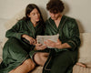 Matching dark green satin robes couple reading a newspaper on a cozy bed.