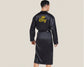 A male model wearing a black satin robe with "King" embroidered in gold along with a crown design on the back. The robe has a smooth, luxurious finish with a tie belt.