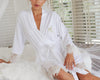 Woman sitting on a fluffy white bed wearing a white robe with lace trim, embroidered with a gold initial "A" on the chest.