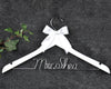 White hanger with "Mrs. Shea" in silver wire, adorned with a white satin bow, set against a dark fabric background with green ivy leaves.