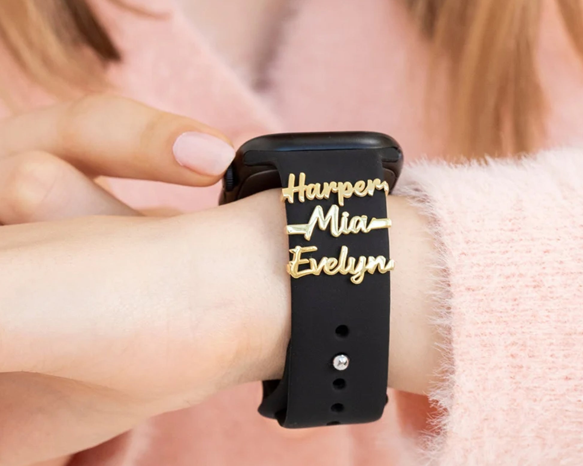 Gold name charms with heartbeat design on a black Apple Watch band, featuring the names Harper, Mia, and Evelyn.