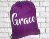 A purple drawstring bag with the name "Grace" written in white cursive lettering, placed against a white brick background.