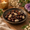 A ceramic bowl filled with polished stones, some dark reddish-brown with gold runic symbols and others light beige, set on a wooden surface with a lit candle, dried flowers, and a purple crystal cluster in the background.