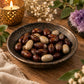 A ceramic bowl filled with polished stones, some dark reddish-brown with gold runic symbols and others light beige, set on a wooden surface with a lit candle, dried flowers, and a purple crystal cluster in the background.