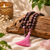 A beaded necklace with pink and dark purple marbled beads and a bright pink tassel, draped over a rustic wood log on a beige fabric surface with decorative flowers, a candle, and green leaves.