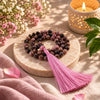 A beaded tassel necklace made of pink and dark marbled beads with a long pink tassel, displayed on a round stone tray with flowers, a lit candle, and green leaves in the background.