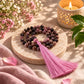 A beaded tassel necklace made of pink and dark marbled beads with a long pink tassel, displayed on a round stone tray with flowers, a lit candle, and green leaves in the background.