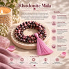 A set of brown and pink rhodonite mala beads with a pink tassel, arranged on a wooden tray over pink fabric, with a lit candle in a glass holder. The image includes text overlays detailing the benefits and product details of the mala beads.