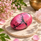 A polished pink stone egg with black veining sits on a round beige stone coaster, surrounded by pink flowers, green leaves, and soft natural light.