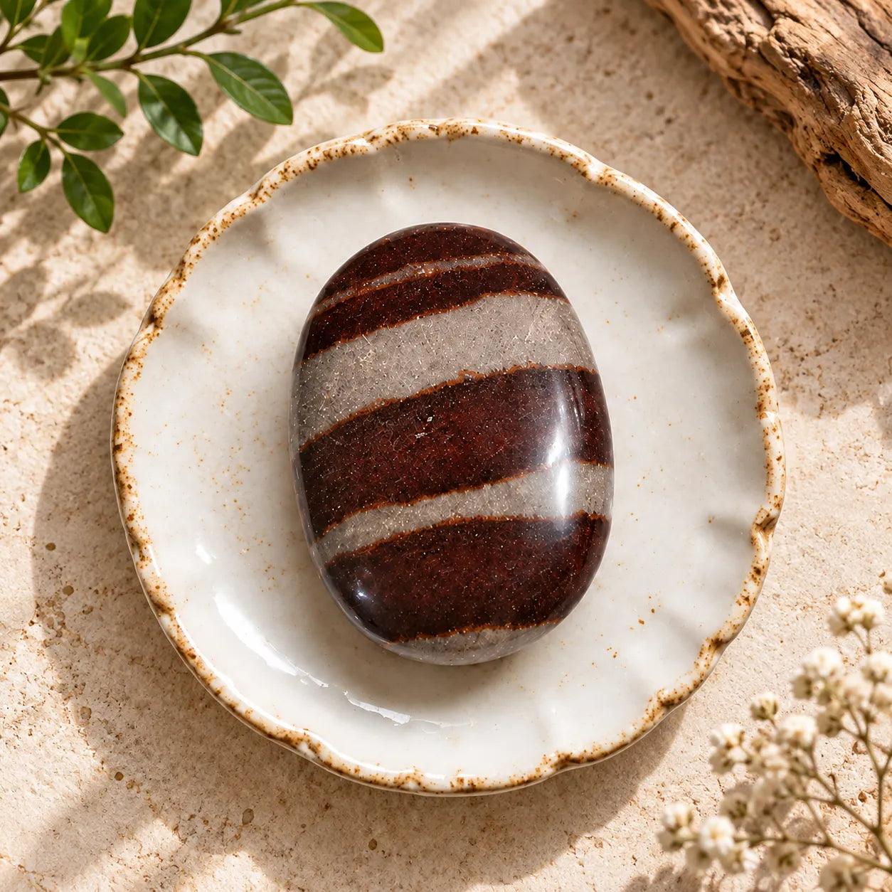 A polished oval stone with dark reddish-brown and light beige horizontal bands sits centered in a small white ceramic dish with a rustic beige rim, styled on a sandy surface with leaves, driftwood, and small white flowers.