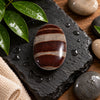 A polished oval stone with horizontal brown and beige bands sits on a dark wet slate slab, surrounded by green water droplets, smooth river stones, and a folded beige towel on a wooden surface.