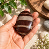 A polished oval stone with alternating dark brown and light beige bands is held in a person's hand, surrounded by decorative stones, green leaves, and small white flowers on a light neutral surface.