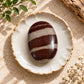 A polished oval stone with dark reddish-brown and light beige horizontal bands sits centered in a small white ceramic dish with a rustic beige rim, styled on a sandy surface with leaves, driftwood, and small white flowers.