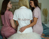 Three women wearing personalized satin bridal pajama sets in blush and white, sitting on a bed with soft decor.