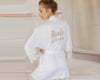 Bride wearing a white lace-trimmed robe with gold embroidery on the back, featuring "Bride" and a wedding date.