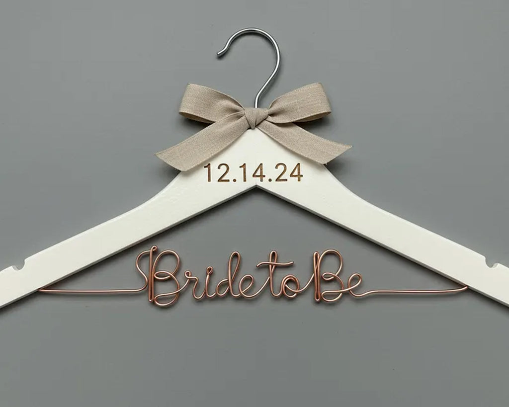 A white wooden hanger with a beige ribbon tied into a bow at the top. The date "12.14.24" is engraved on the hanger, and the phrase "Bride to Be" is crafted in elegant copper wire below.