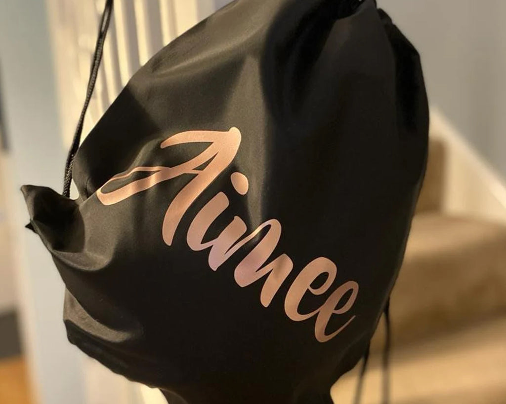 A black drawstring bag with the name "Aimee" written in elegant, cursive, rose gold lettering, hanging on a staircase railing.