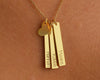 A close-up of a gold necklace featuring three vertical bar pendants engraved with the names "Hannah," "Robbie," and "Maxine," along with a small gold heart charm, worn against the skin.
