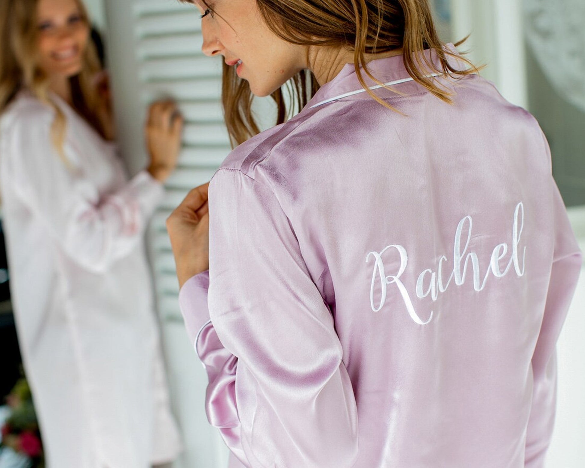 A woman wearing a lilac satin nightshirt with "Rachel" elegantly embroidered on the back, while another woman in a similar outfit smiles in the background.