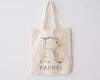 Beige canvas tote bag with the letter "R" beautifully designed with floral and greenery elements, with "RACHEL" printed below, hanging against a white background.