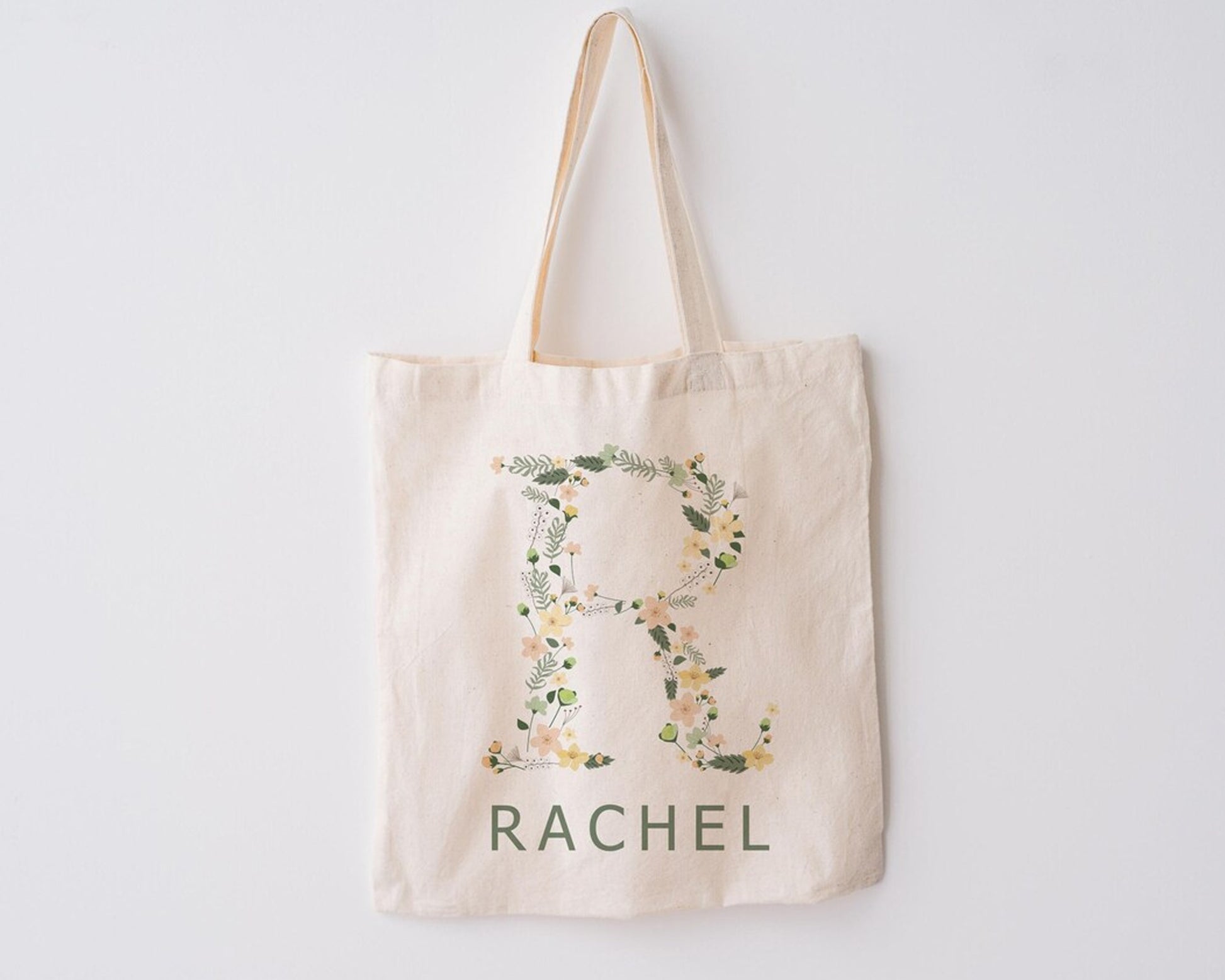 Beige canvas tote bag with the letter "R" beautifully designed with floral and greenery elements, with "RACHEL" printed below, hanging against a white background.