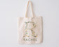 Beige canvas tote bag with the letter "R" beautifully designed with floral and greenery elements, with "RACHEL" printed below, hanging against a white background.