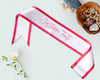 White satin sash with red trim and customizable text, ideal for special events, parties, and celebrations.