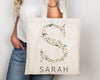 Beige tote bag featuring the letter "S" adorned with delicate green leaves and soft pastel flowers, with "SARAH" written below.