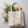 Elegant beige tote bag with the letter "S" formed by delicate green leaves and soft pastel flowers, with "SARAH" printed underneath, surrounded by fresh greenery and orange flowers.
