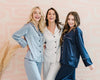 Three women laughing together in silky satin pajama sets in pastel blue, white, and navy, featuring contrast piping and a relaxed fit.