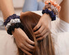 A close-up of two hands adorned with satin scrunchies in various colors, resting on the head of a person with long brown hair.