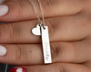 Close-up of a silver bar pendant necklace engraved with "Emily," accompanied by a small heart charm, held in a person's hand.