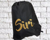 A black drawstring bag with the name "Siri" written in gold glittery cursive lettering, placed against a white brick background.