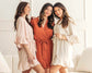 Three women in peach, rust, and cream ruffle robes, smiling in a bright, elegant setting.