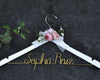 White hanger with "Sophia Rose" in gold wire, decorated with pink and white flowers and a sheer white ribbon, set against a dark fabric background with green ivy leaves.
