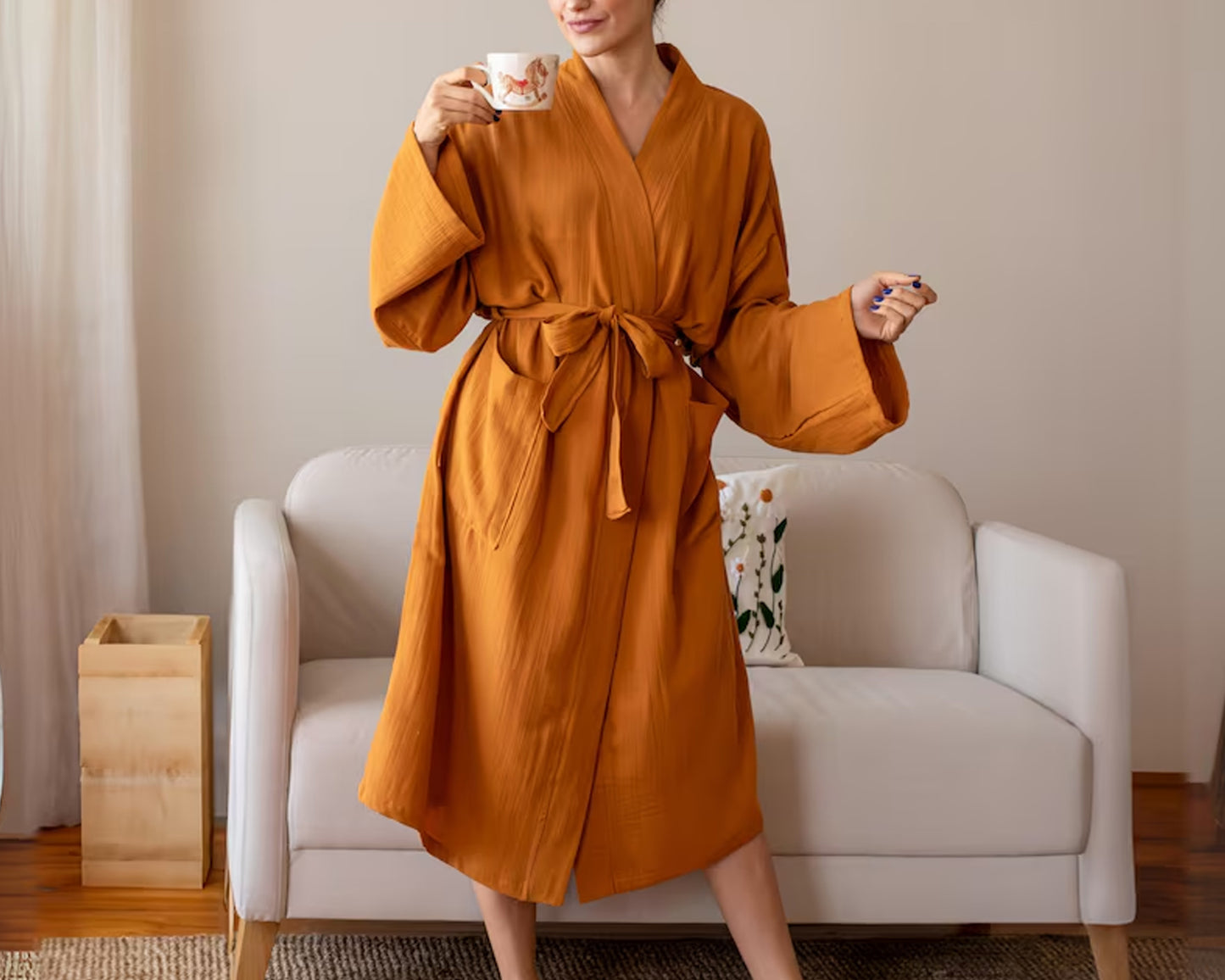 A woman in a burnt-orange belted robe holding a patterned mug, standing in front of a cozy white sofa with decorative pillows.