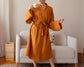 A woman in a burnt-orange belted robe holding a patterned mug, standing in front of a cozy white sofa with decorative pillows.