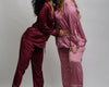 Two women wearing silky satin pajama sets in deep burgundy and soft pink, featuring button-up tops with contrast piping and matching pants.