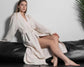 A woman wearing a cream-colored waffle-textured robe, sitting on a black leather couch with crossed legs, against a white wall.