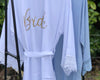 A white bridal robe with lace-trimmed sleeves and a waist tie, featuring "Bride" in gold script on the back, hanging next to a light blue lace-trimmed robe.
