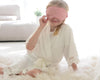 A young girl in a white satin robe with lace trim, playfully holding a pink sleep mask over her eyes while sitting on a fluffy rug.