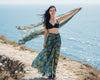 Woman in a black bikini top and patterned sarong standing on a coastal path with a scarf.