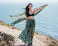 Woman in a black bikini top and patterned sarong standing on a coastal path with a scarf.