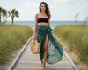 Woman in a black top and patterned sarong standing on a wooden path with grass and ocean in the background